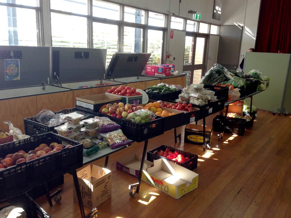 A vibrant display of fresh fruits and vegetables arranged on tables in a well-lit indoor market. The scene features a variety of produce, including tomatoes, apples, and leafy greens.