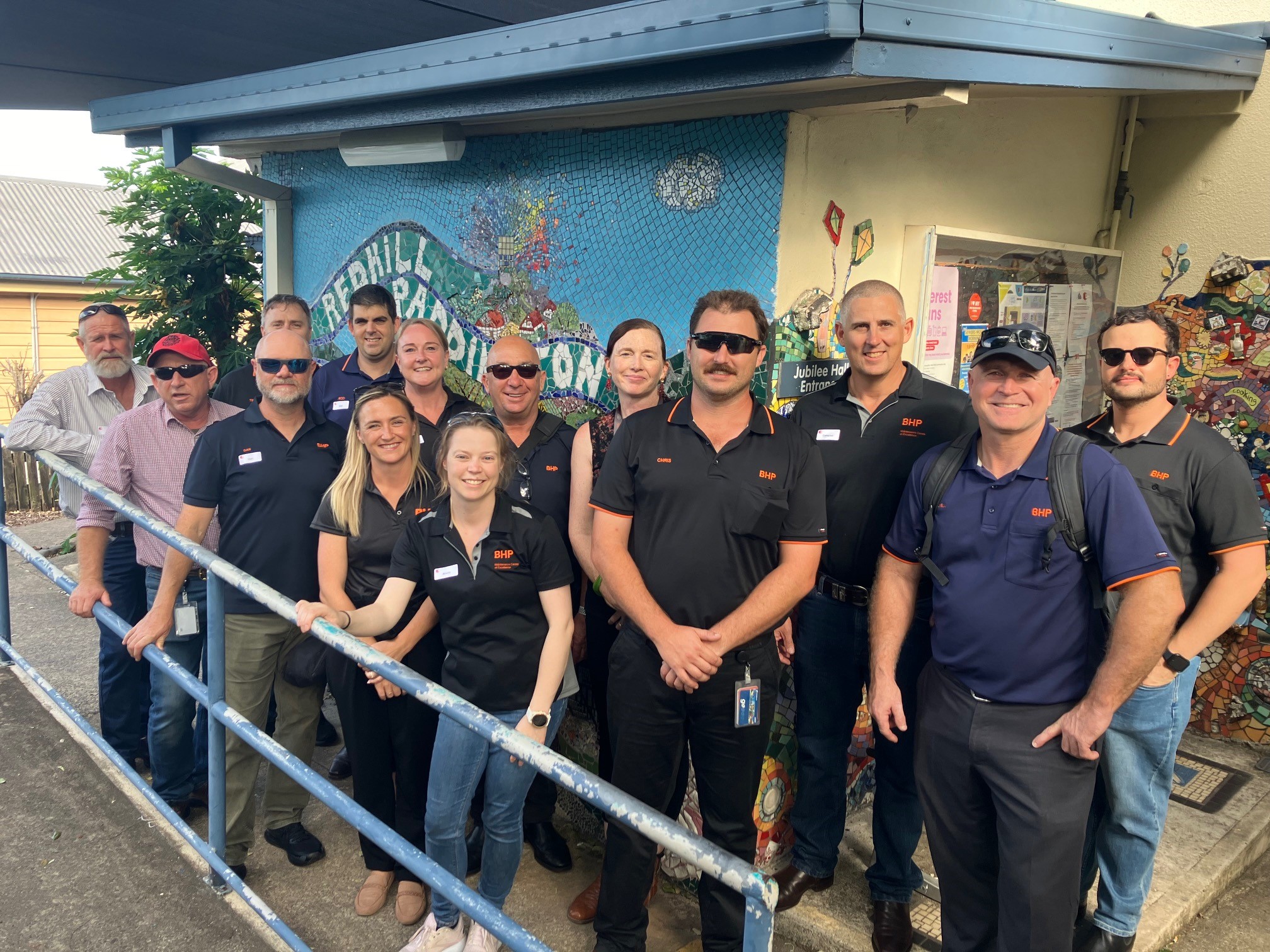 A diverse group of individuals standing together in front of a colorful mural. They are wearing black shirts, with some wearing hats and sunglasses. The group includes both men and women, displaying a mix of ethnicities, all smiling or looking confidently at the camera.