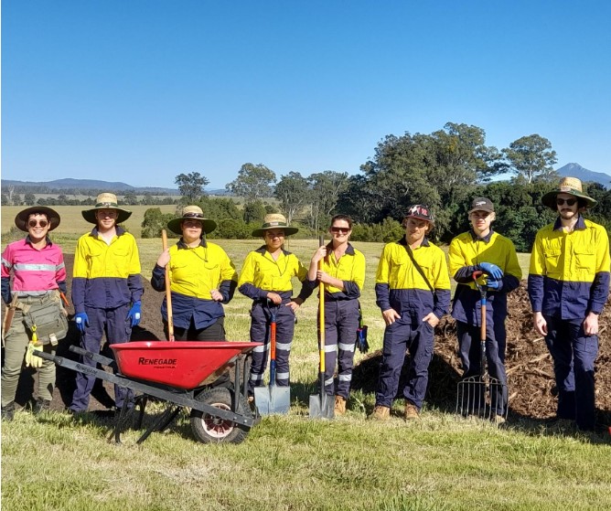 Group of diverse individuals in yellow and navy work uniforms standing in a field, smiling confidently. They are equipped with gardening tools and a red wheelbarrow. The group consists of men and women of varying ethnicities, showcasing teamwork in outdoor landscaping or gardening activities.