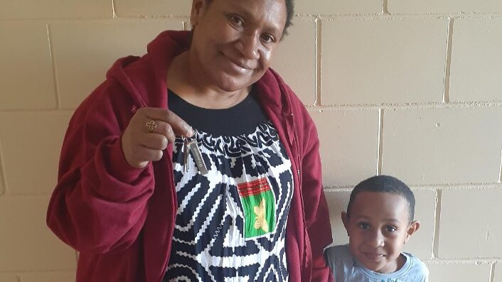 A smiling woman of Papua New Guinean descent holds keys in her right hand, wearing a black and white patterned dress with a colorful emblem, complemented by a red zip-up jacket. Beside her, a young boy wears a light blue shirt and smiles at the camera.