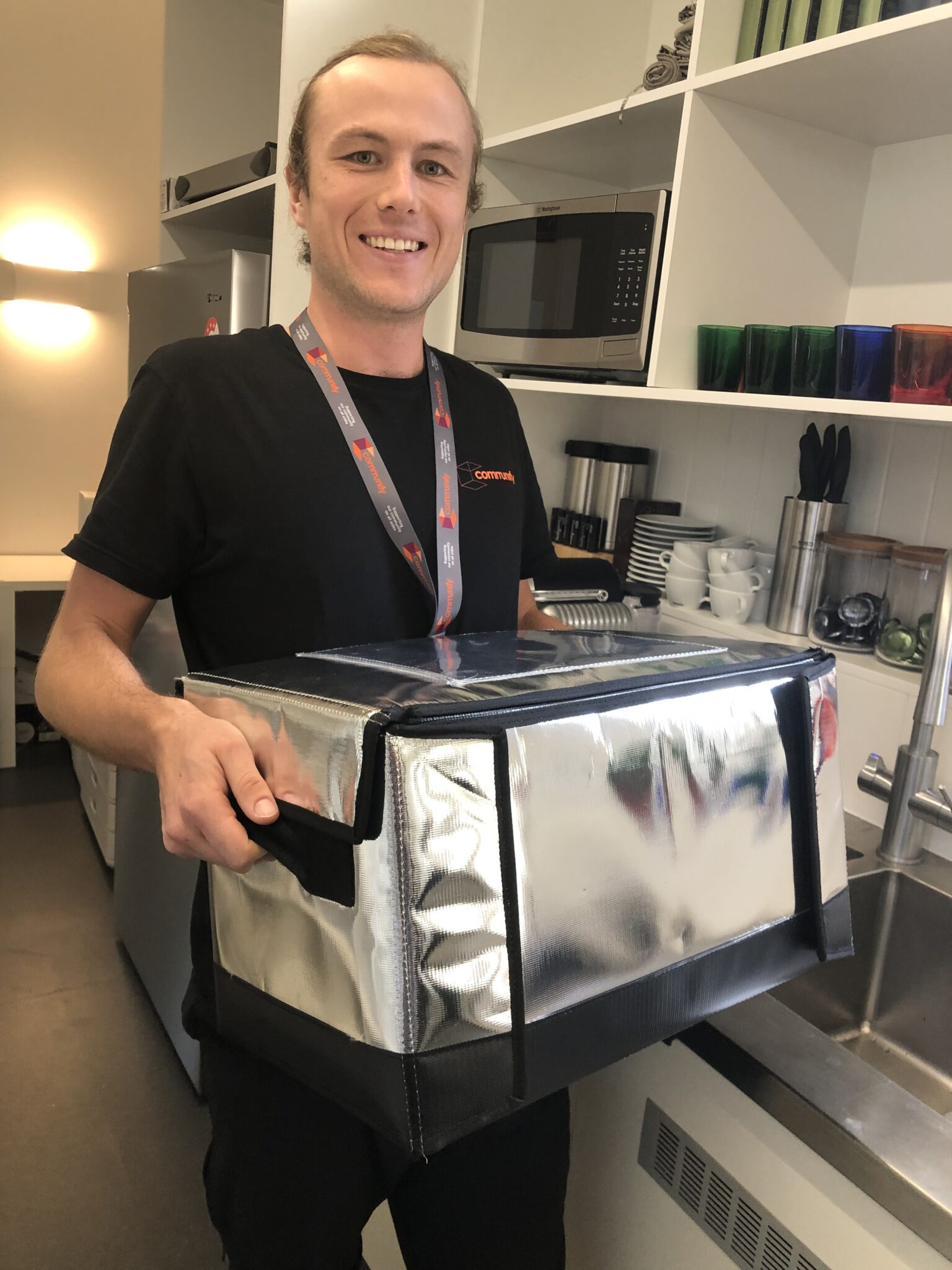 A smiling individual with light skin and short brown hair holds a silver insulated food delivery box in a modern kitchen setting. They are wearing a black shirt and a lanyard around their neck.