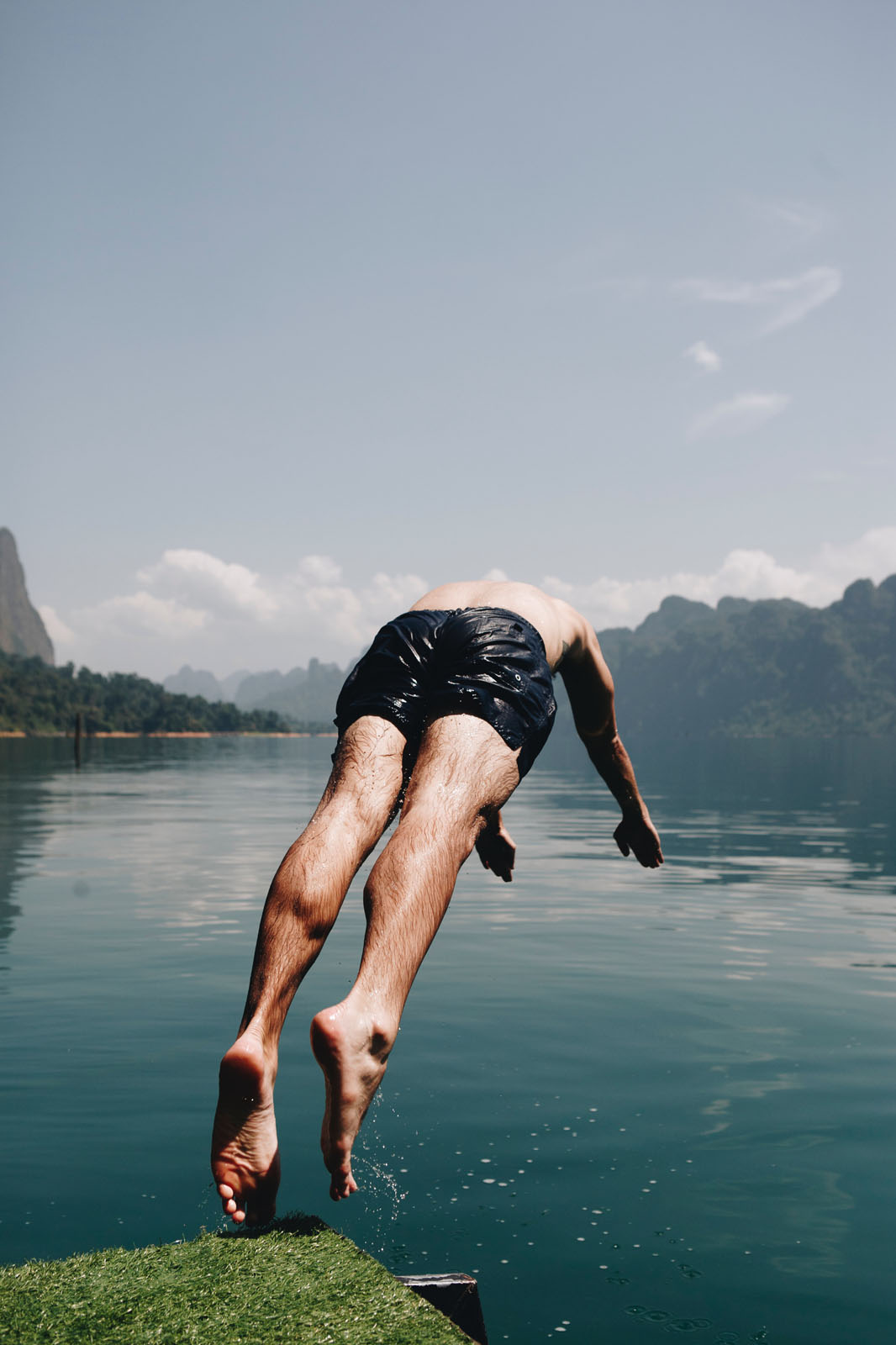 A person diving into a lake from a rocky ledge, wearing black swim shorts. The person is captured mid-air, diving into the water. Surrounding landscape includes mountains and trees in the background.