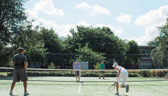 A group of three men playing tennis on an outdoor court. One man, wearing a white shirt and shorts, is preparing to hit the ball, showing concentration. The second man, dressed in a blue shirt and dark shorts, stands ready to receive the ball, while the third man, in a green shirt and light shorts, watches from the side. The background features lush trees surrounding the court.