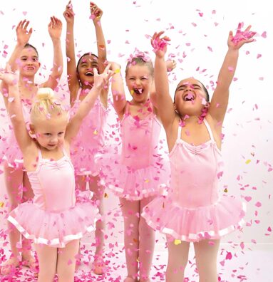 A group of five young girls celebrating in pink tutus and leotards, displaying joyful expressions. The girls, of various ethnicities, are throwing pink confetti in the air, with their arms raised in excitement.