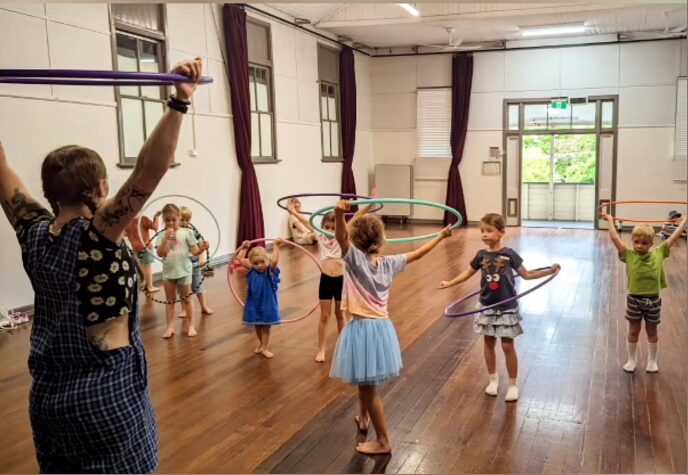 Children participating in a hula hoop activity in a spacious room. A teacher, wearing a black top with white polka dots and blue checkered pants, demonstrates the activity. The children, of diverse ethnicities, are dressed in colorful outfits, including a light blue dress, a white shirt with stripes, and a pink skirt. They display expressions of joy and concentration while engaging with their hula hoops.