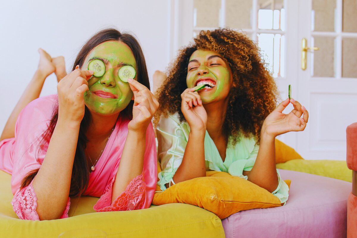 Two women enjoying a skincare routine at home. The white woman with straight dark hair wears a pink robe and holds cucumber slices over her eyes, smiling playfully. The black woman has curly hair, wears a light green robe, and is laughing while holding a small jar of skincare product. Both have green facial masks applied.